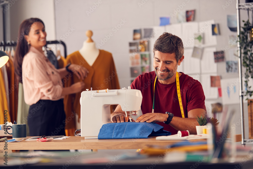 Fototapeta premium Male Fashion Designer Working On Sewing Machine As Female Colleague Works In Background