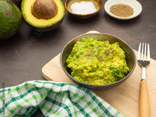 Fresh guacamole on a dish placed on a cutting wooden board with ingredients for homemade guacamole: avocados, lemon, salt, and pepper. Top view. Concept of traditional Mexican preparation