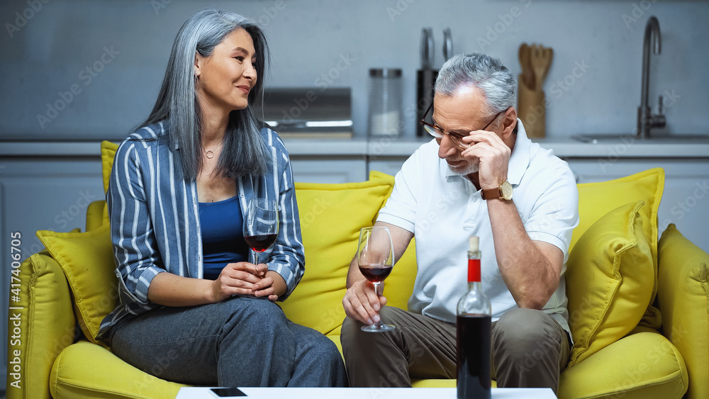 happy elderly interracial couple holding glasses with red wine on sofa at home happy elderly interracial couple holding glasses with red wine on sofa at home