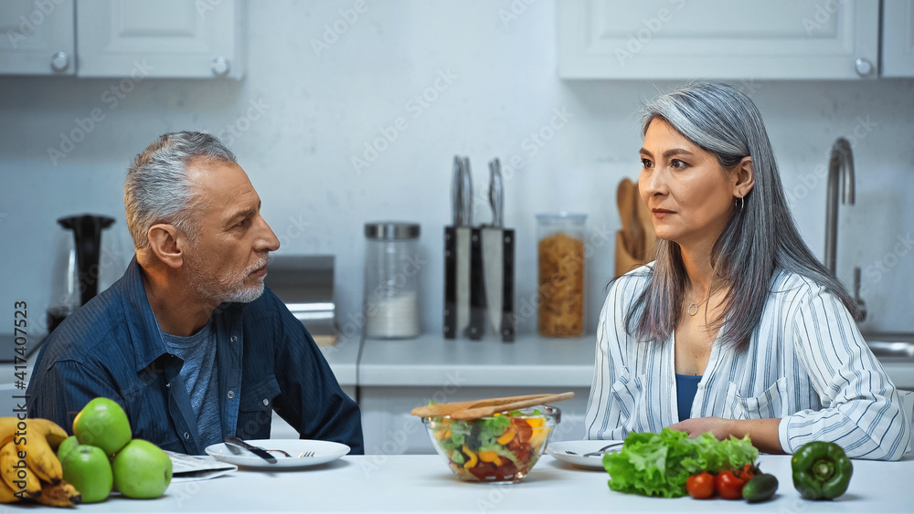 elderly interracial couple looking at each other during breakfast in kitchen