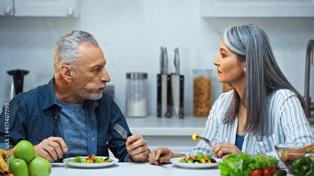 senior interracial couple talking while eating fresh salad in kitchen