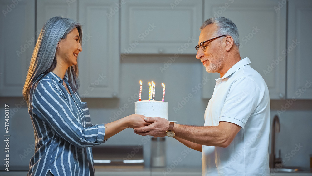 side view of pleased senior man taking birthday cake from happy asian wife