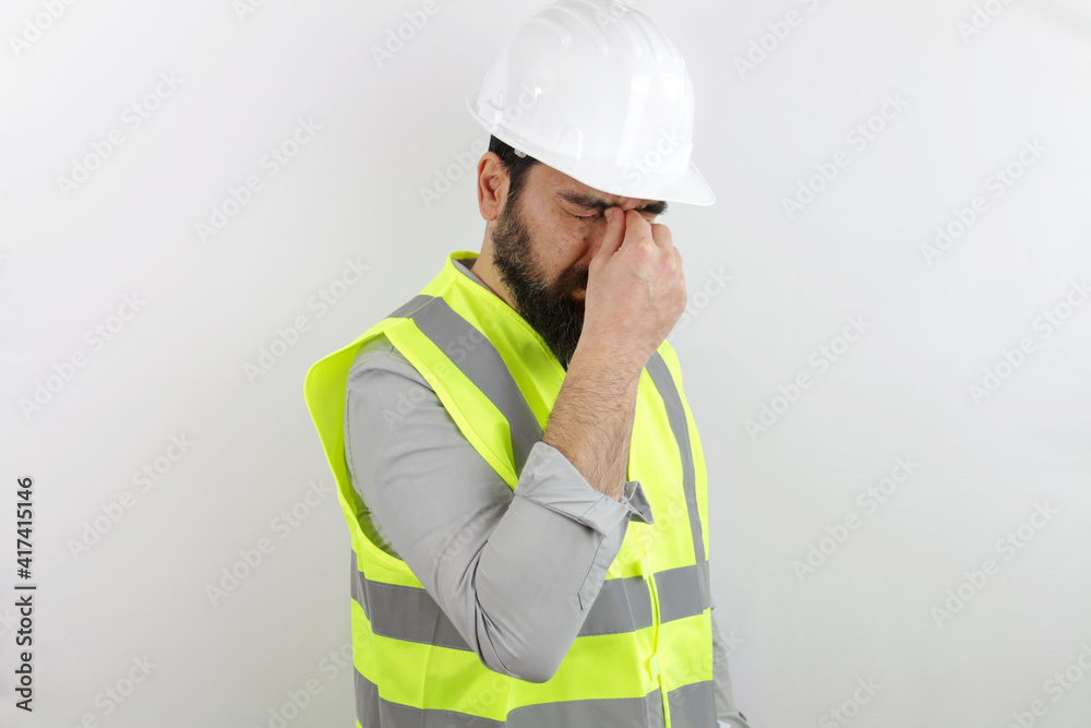 Bearded man architect wearing safety helmet and reflective jacket tired rubbing nose and eyes feeling fatigue and headache. Stress and frustration concept.