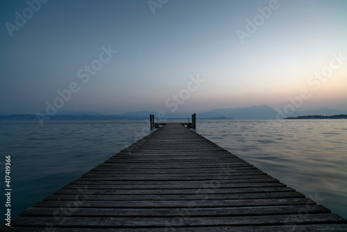 Fototapeta Naklejka Na Ścianę i Meble -  An old wooden pier extends into the clear blue waters of the lake. A solitary path towards the calm and peaceful silence of nature, with the sweet sound of the waves.
