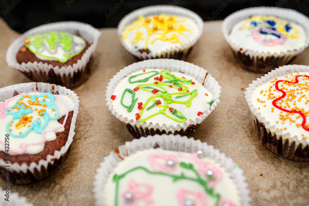 Chocolate brown muffins wrapped in white paper and covered with white frosting with colorful decorations, baked in the oven, lying on baking paper.
