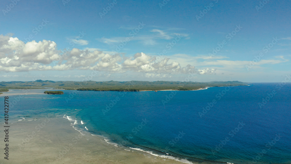 Coast of Siargao island is covered with forests and waves crashing on a coral reef. Seascape: ocean with blue water against the sky and clouds.