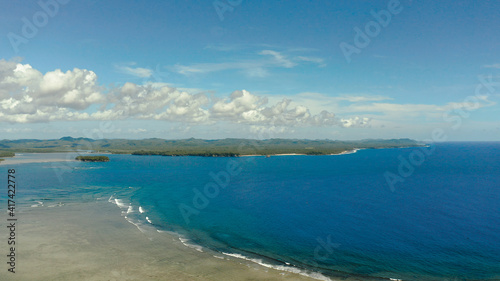 Coast of Siargao island is covered with forests and waves crashing on a coral reef. Seascape: ocean with blue water against the sky and clouds.