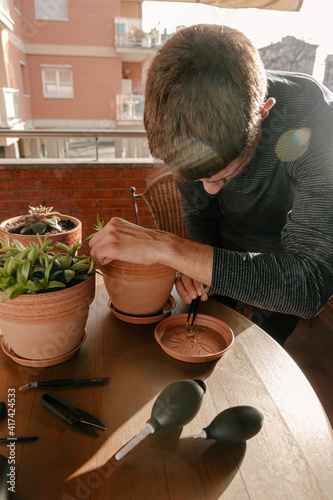 Hombre joven caucásico cuidando varias de sus plantas en la terraza con variedad de herramientas en la mesa de su casa durante el atardecer.