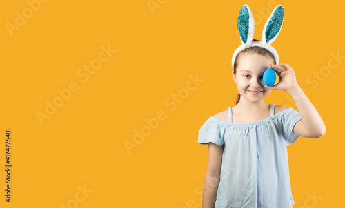 Happy Easter, kids. A girl in rabbit ears on her head holds a blue egg in her hands and closes one eye with it. Baner,