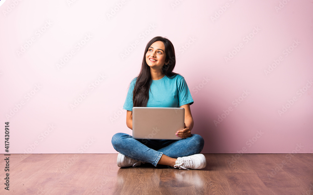 Indian asian girl using laptop while sitting against pink wall on ...