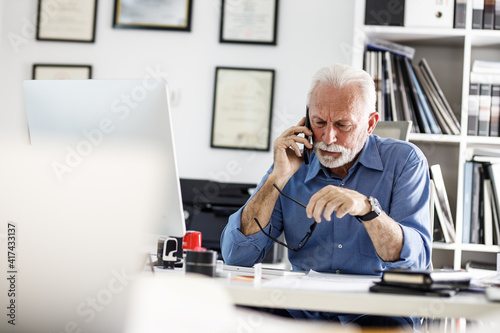 Senior business manager sitting at the desk in his office and speaking on smart phone.Looking serious and trying to solve problem.