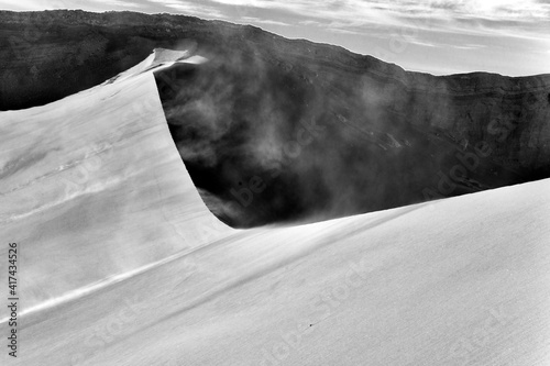 Black and white image of a sand dune, wind blowing sand over the ridge, and a rocky cliff in the background, Valle de la Luna, Atacama desert, Chile
