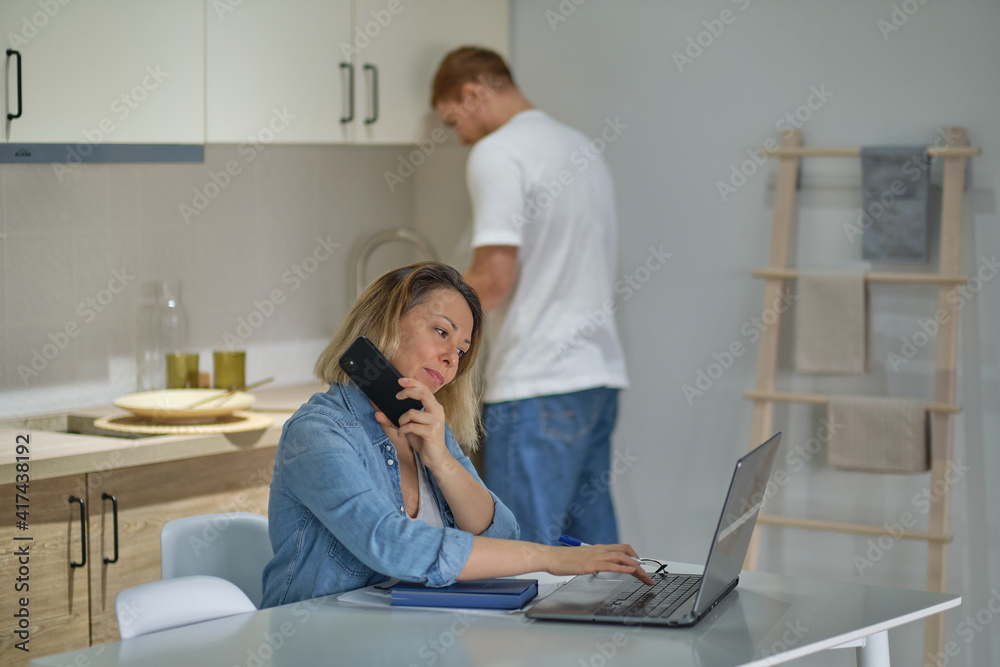 Obraz premium busy business woman talking on the phone and using the computer at home in the kitchen. the partner or husband in the background is cooking. healthy relationships modern multicultural family