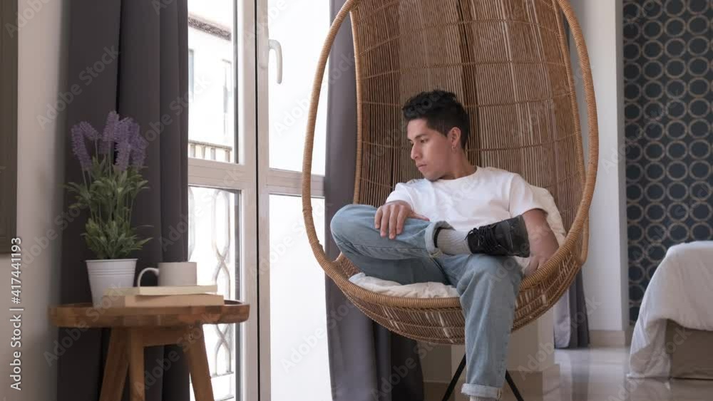 young man looking out the window sitting in a hanging chair at home young man looking out the window sitting in a hanging chair at home