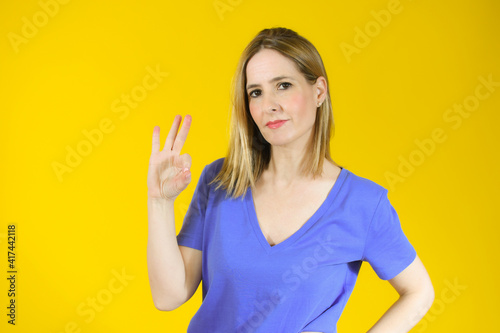 Young woman standing over yellow background smiling positive doing okay sign with hand and fingers. Successful expression.