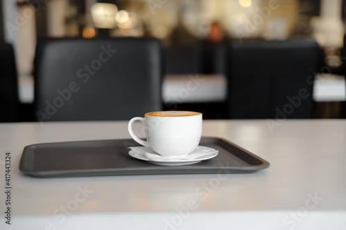 a white cup of coffee with a saucer on a black tray on a white table against the background of lights in a cafe with a black and white interior