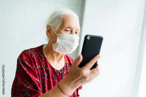 An old woman is talking on the phone standing at the window in a medical mask. Quarantine, stay at home, coronavirus infection, covid-19