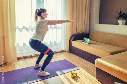 Young attractive woman in sportswear doing workout at home, doing sit-ups squats with rubber resistance band on floor violet mat and watching online tutorials video on tablet screen in living room