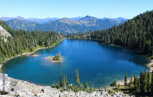 Top view of a lake with a small island surrounded by pine trees, mountains and rocks