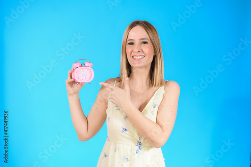 Young woman with blue eyes holding alarm clock standing over isolated yellow background very happy pointing with hand and finger