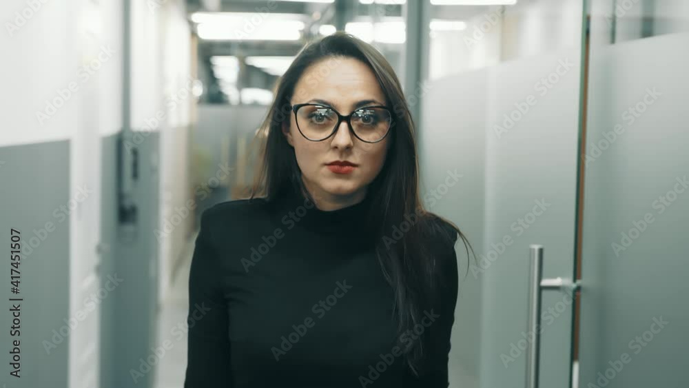 A woman in glasses and black clothes walks down the hall and looks into the camera. A woman in glasses and black clothes walks down the hall and looks into the camera.