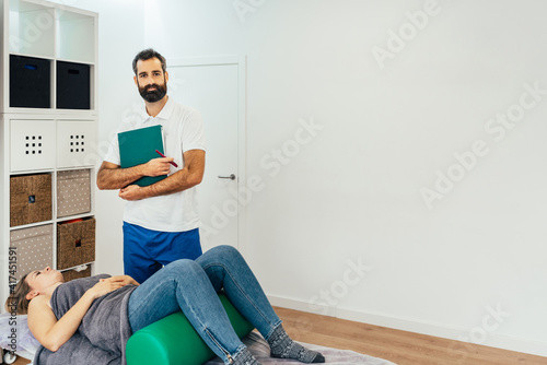 Portrait of a standing masseur holding a clipboard in his clinic. The doctor with beard and friendly gesture looking at the camera.