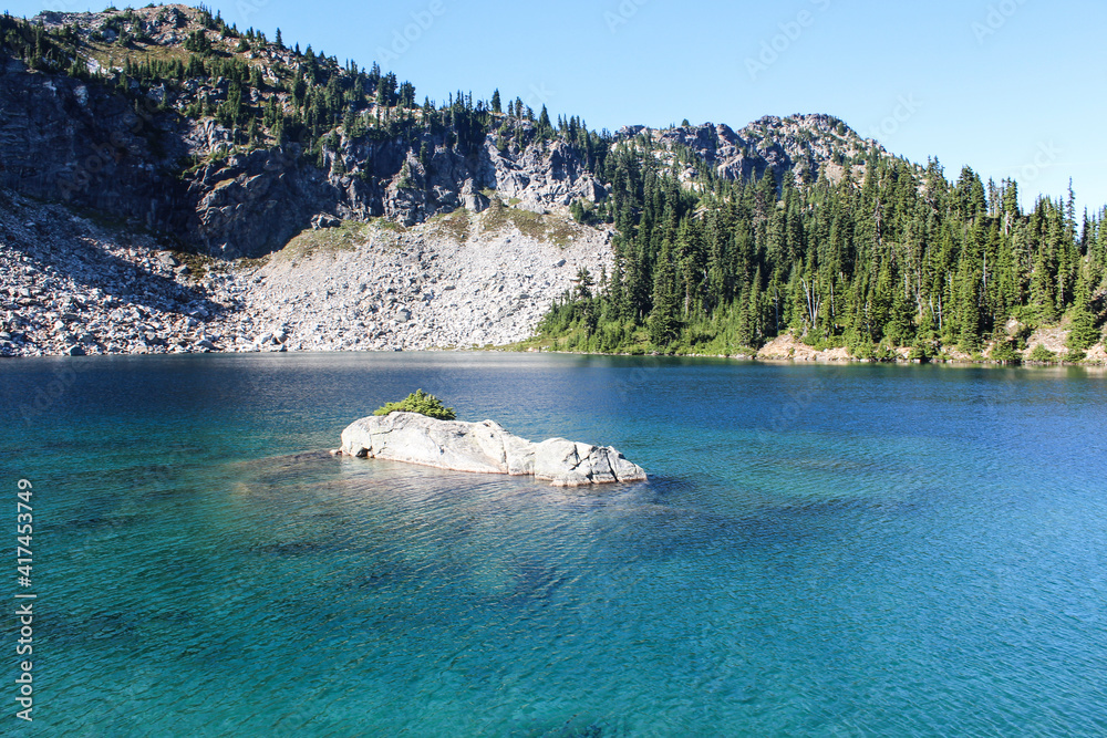 Naklejka premium beautiful crystal clear aquamarine lake with a big rock in the middle surrounded by mountains, pine trees and rocks