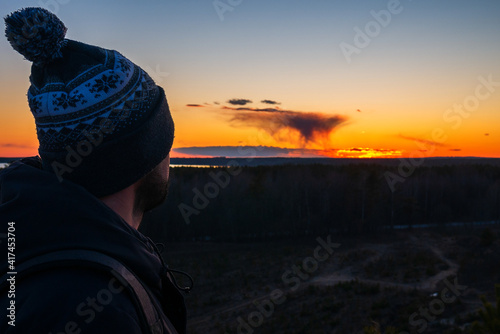 A man in a hat with a backpack stands on top of a mountain against the backdrop of a red sunset. A lonely man in the background of the sun. Face in profile.