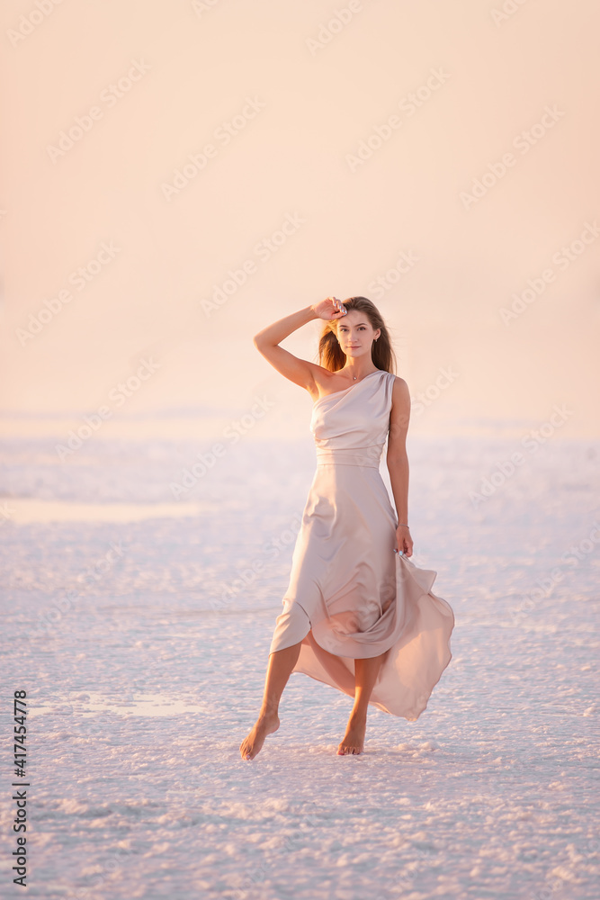 Young blonde woman in an evening airy pastel pink, powdery dress stands barefoot on white crystallized salt. Girl with natural make-up, hair is developing. Salt mining trip, walking on water at sunset Young blonde woman in an evening airy pastel pink, powdery dress stands barefoot on white crystallized salt. Girl with natural make-up, hair is developing. Salt mining trip, walking on water at sunset