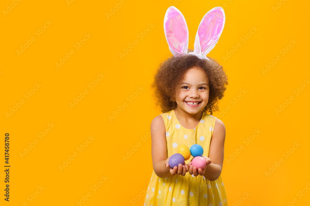 An african girl in sunny orange dress with rabbit ears on her head with painted eggs in her hands on a yellow background