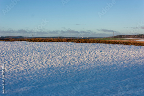  Landscape of the end of winter. In the foreground is a snow-covered part of the field with blue shadows. In the distance - a part of the field free of snow and a forest lit by the morning sun.