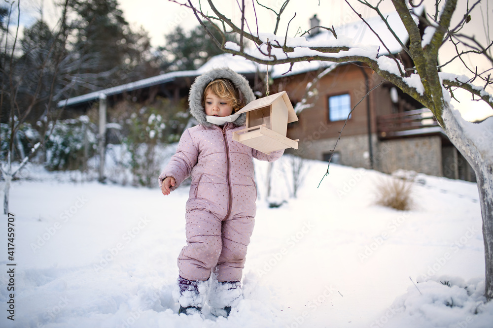 Small girl outdoors in winter garden, standing by wooden bird feeder.