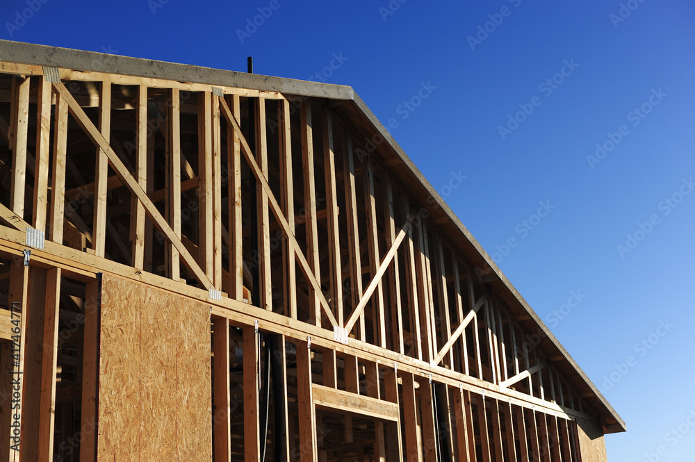 Obraz premium Close-up of gable-end framing with vertical studs, angled roof trusses, and partial sheathing on a residential building under construction against clear blue sky