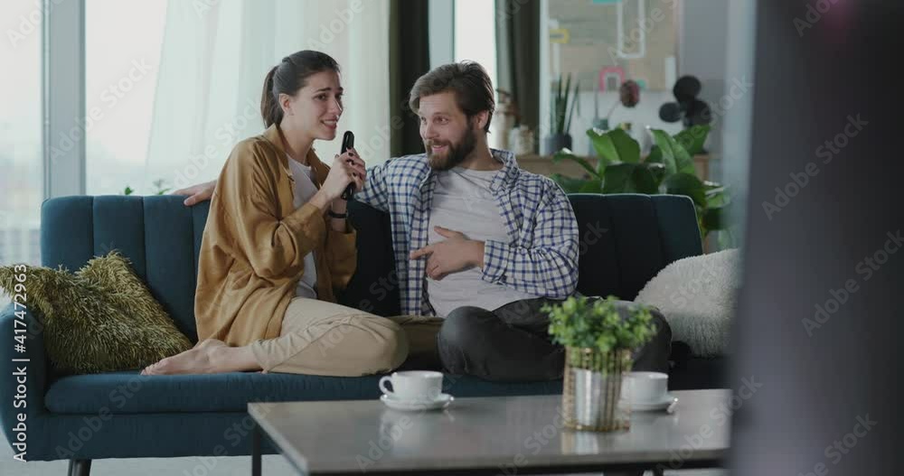 Young caucasian couple stay home watching TV. Funny excited woman using remote control as microphone singing karaoke making boyfriend smile. Young caucasian couple stay home watching TV. Funny excited woman using remote control as microphone singing karaoke making boyfriend smile.