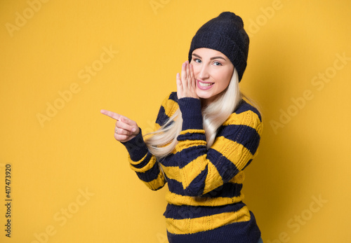 Woman holding hand near mouth and telling secret, model wearing woolen cap and sweater, isolated on yellow background. Whispering concept photo