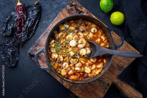 Traditional slow cooked Mexican pozole rojo soup with ground minced beef, hominy maiz and dry ancho paprika as top view in cast-iron roasting dish on old rustic wooden board photo