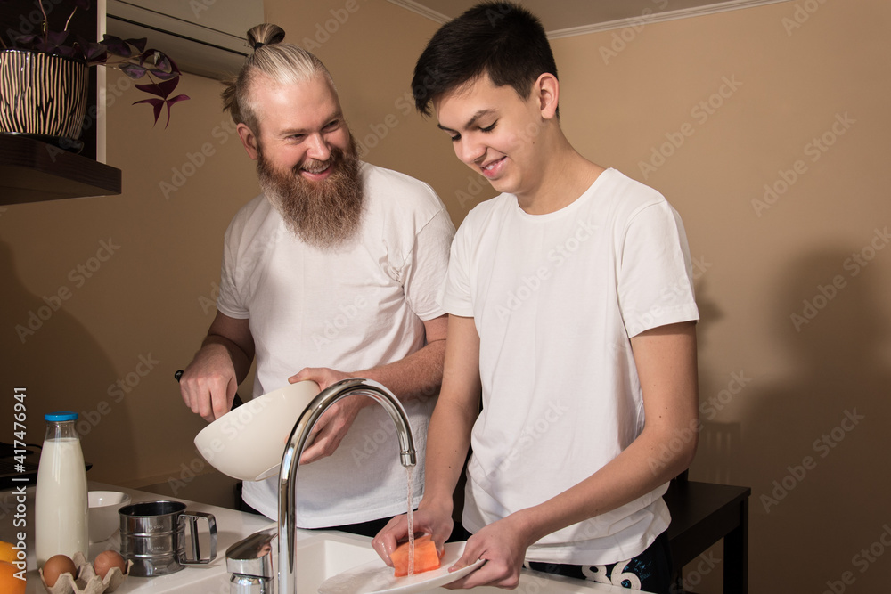 Happy dad and son teenager on kitchen. Smiling father cooking, son washing the dishes. Concept of friendly relationship between parents and children teens.