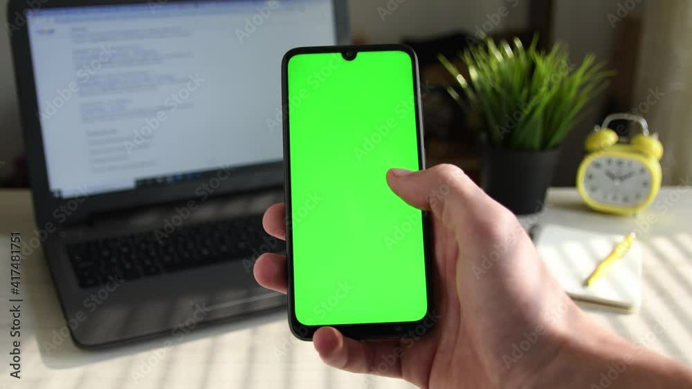 Young man holds a mobile smartphone in his hand and flips swipes right his finger on the screen on the background of the desktop, laptop