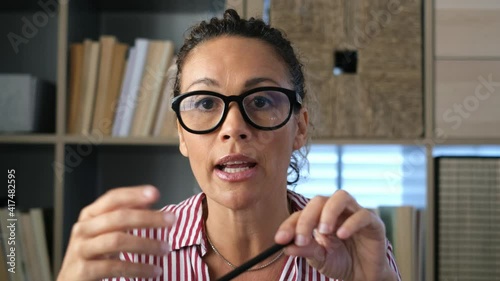Head shot portrait confident businesswoman coach wearing glasses looking at camera and talking, mentor speaker holding online lesson, explaining, sitting at wooden work desk in modern cabinet
