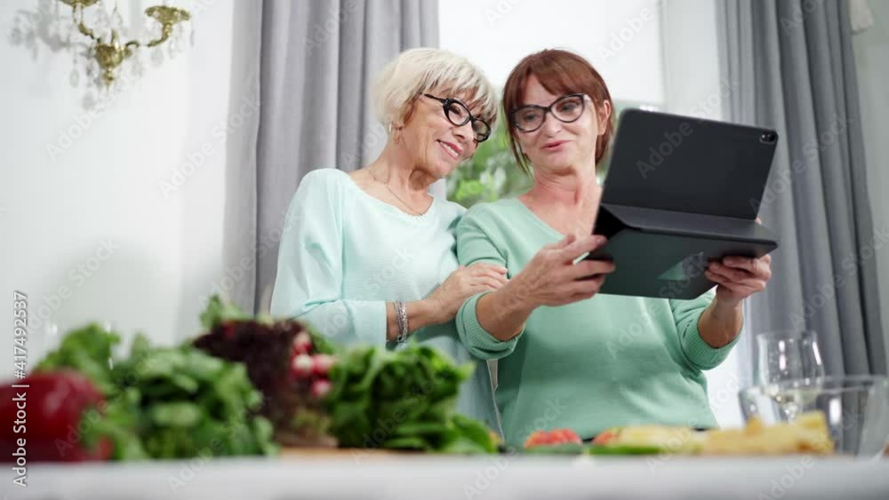 Middle shot portrait of Caucasian retirees in eyeglasses discussing online news holding tablet and talking. Beautiful senior women gossiping and cooking healthful salad indoors. Middle shot portrait of Caucasian retirees in eyeglasses discussing online news holding tablet and talking. Beautiful senior women gossiping and cooking healthful salad indoors.