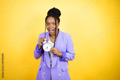 Young african american business woman surprised holding and pointing a clock