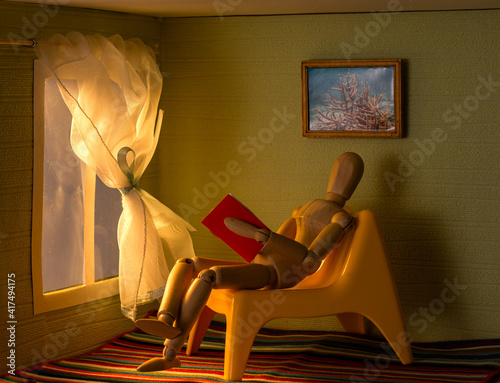 Figurine of a puppet man sitting on an armchair in a toy room and reading a book by the window at sunset