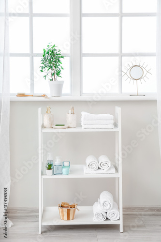 Shelf unit with cosmetics and towels near window in bathroom