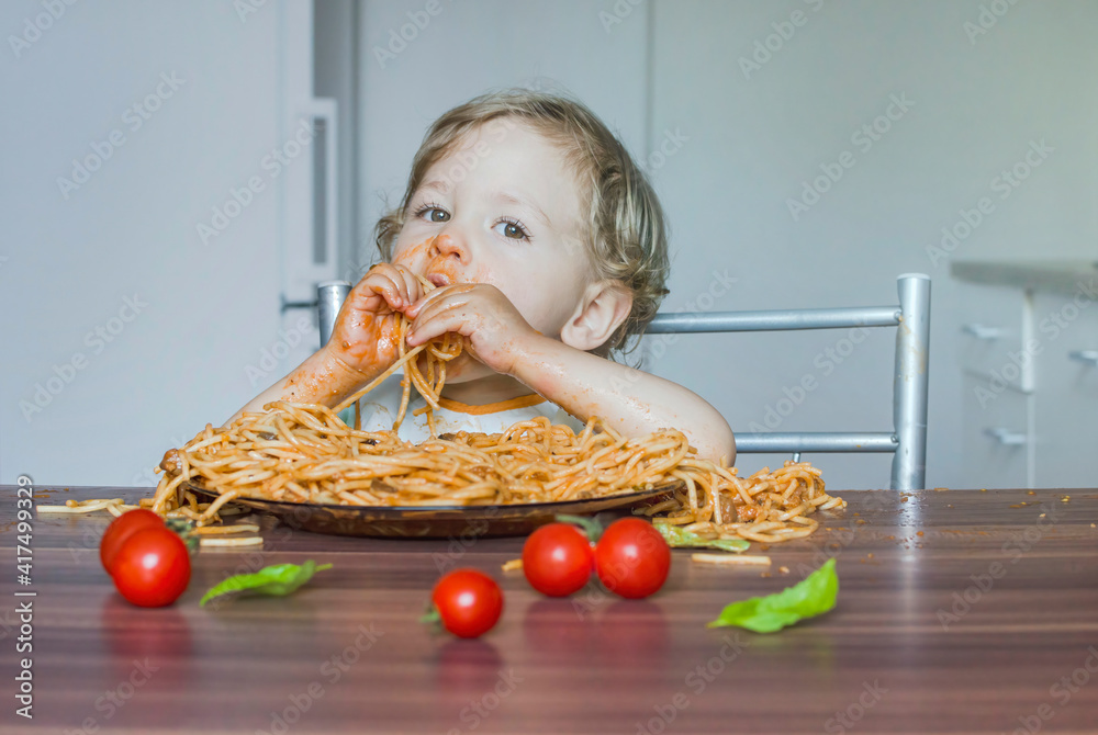 Funny baby child getting messy eating spaghetti with tomato sauce from a large plate, by itself with his hands, at home Funny baby child getting messy eating spaghetti with tomato sauce from a large plate, by itself with his hands, at home