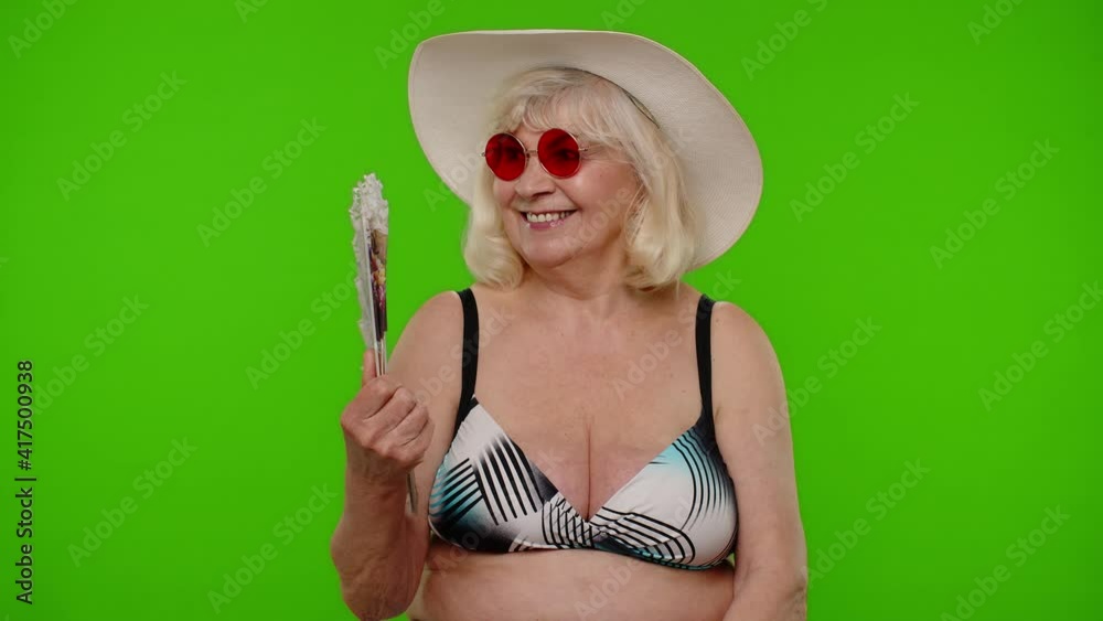 Senior pensioner woman tourist exhales from heat or stuffiness, waves hand fan at herself on chroma key background. Portrait of exited happy old granny traveler in swimsuit model on summer vacations