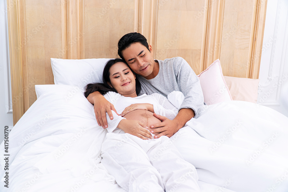 Fototapeta premium Pregnancy, parenthood or family concept. Cheerful handsome young man embrace his pregnant wife touching on tummy while lying relax togetherness on the white bed.