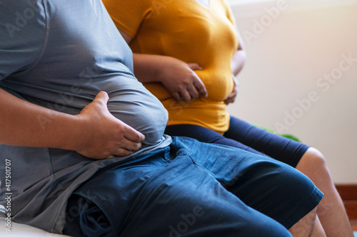 Фотография Fat man and woman holding their big belly while sitting on the bed suffering from extra weight