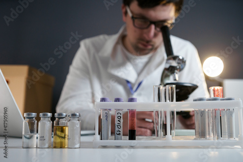 Scientist in lab coat and eyeglasses working at workplace with laptop in laboratory. Male doctor researcher are doing investigations with microscope and laptop. Microbiologists in laboratory