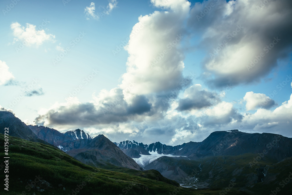Dramatic alpine landscape with mountain ridge under cloudy sky. Large cloud in blue sky above silhouettes of mountain range. Big cloud in gradient sky above mountains silhouettes and beautiful glacier