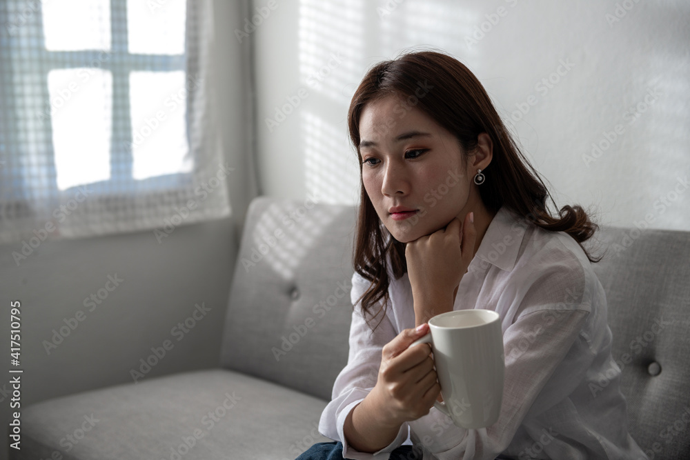 Unemployed Asian woman at home alone on a sofa with a cup of coffee thinking for a new job and has no money because the company closed. Lose a job during Coronavirus or Covid-19 quarantine concept